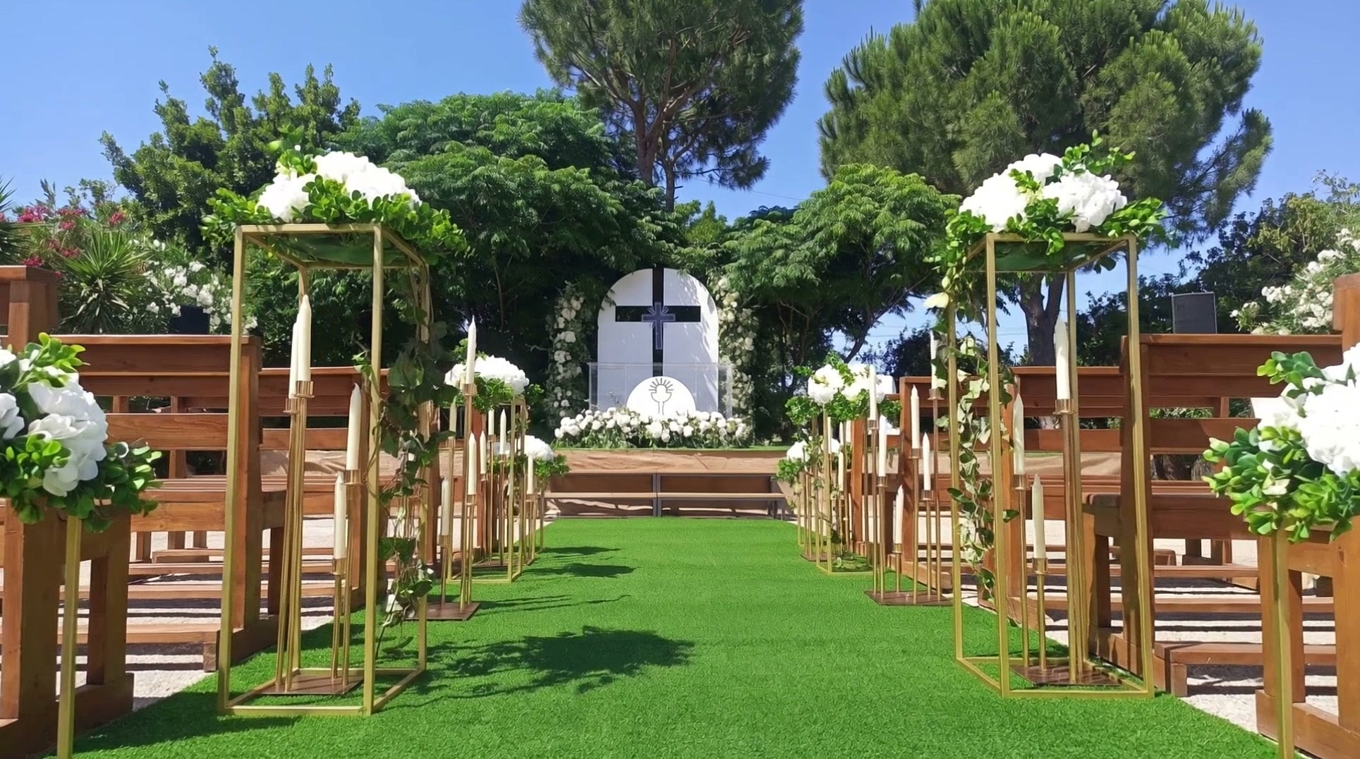 Outdoor ceremony aisle lined with gold candle stands and white hydrangeas, leading to a white altar with a cross