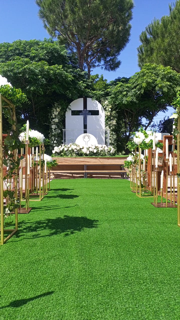 Outdoor ceremony aisle on a lawn, lined with gold stands and white flowers, leading to a cross backdrop